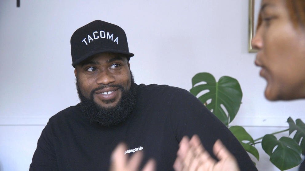 A man in a black “Tacoma” hat smiles as a woman speaks to him
