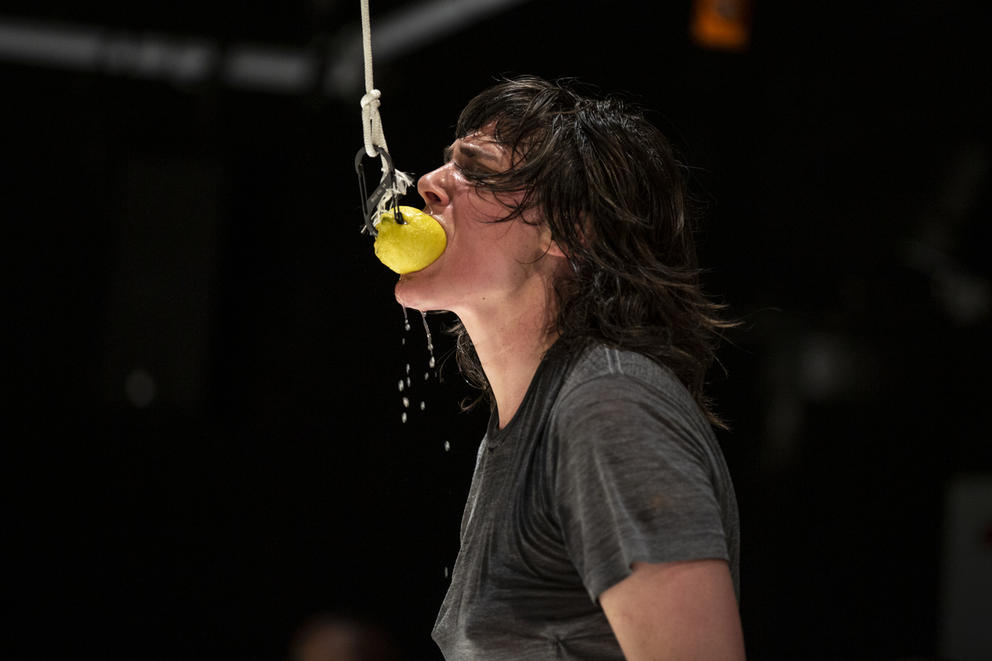 Person in gray t-shirt on black background bites in a lemon suspended from the ceiling