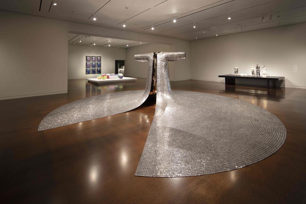 Museum gallery featuring, in the middle, a large sculpture that looks like a robe made from metal, its long sleeve extending over the floor