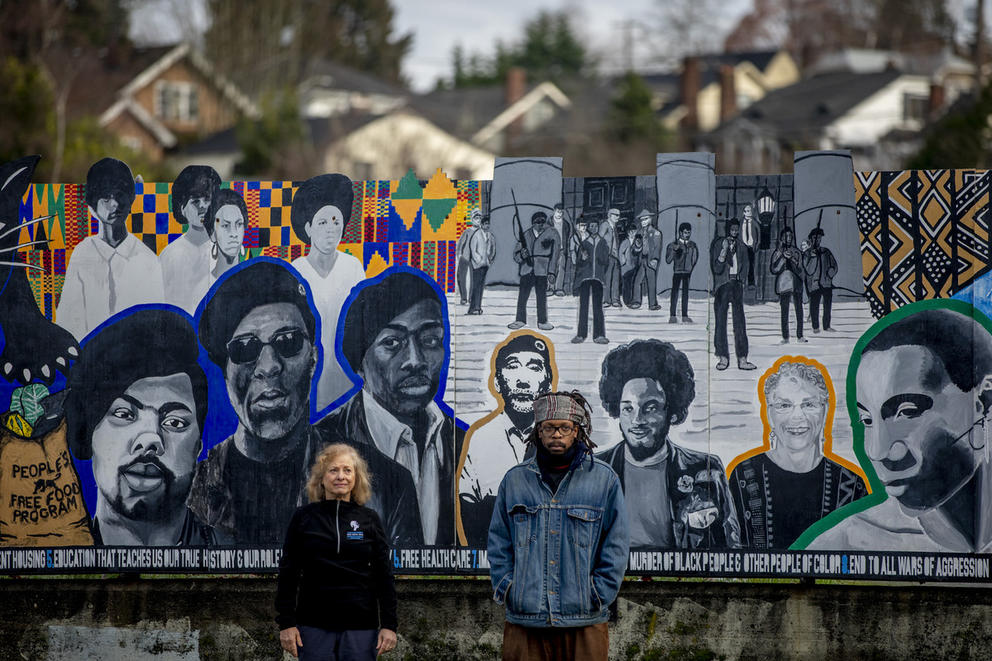 A woman and man standing in front of a Black Panther Party mural depicting civil rights heroes
