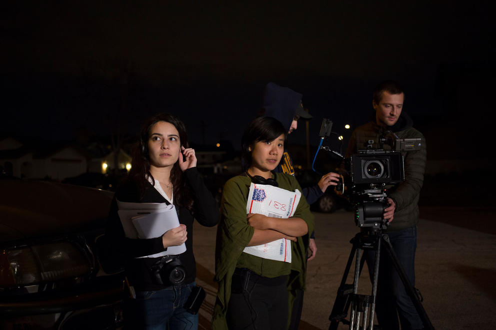 Two people on a film set, one holding stack of paper. A person with camera in the background.