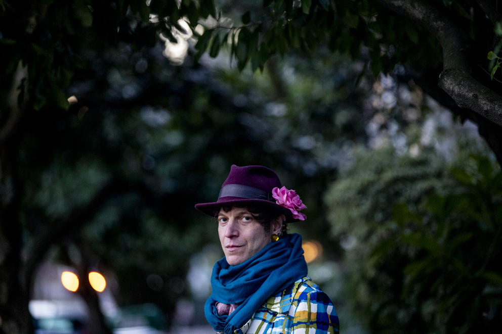 Person with blue scarf, plaid jacket and purple hat with pink flower amid the trees