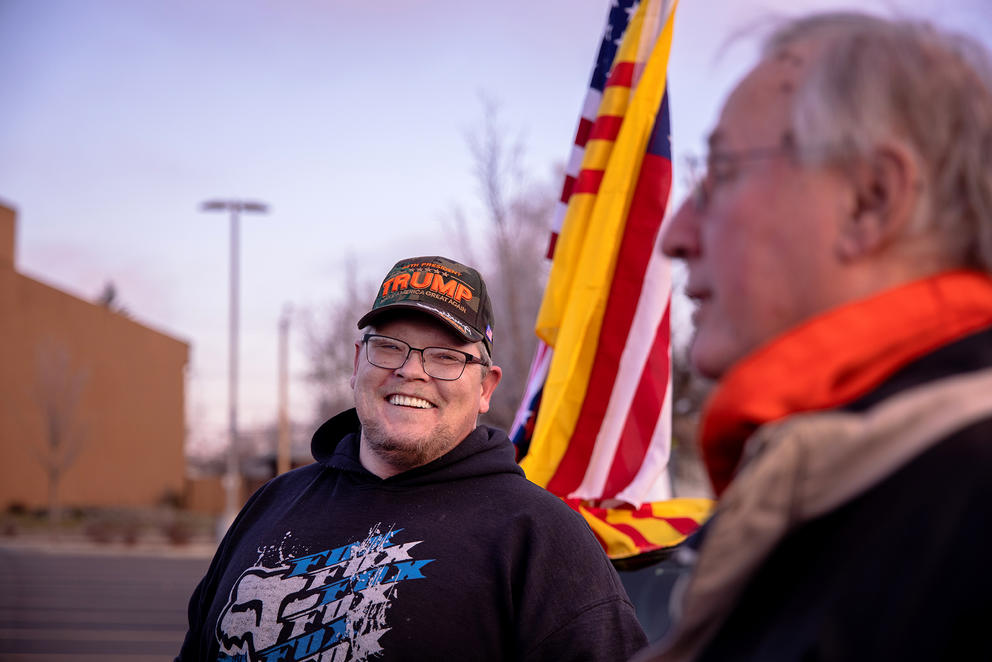A man smiles outside with a flag behind him
