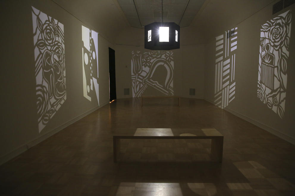 Exhibit room at the Henry art gallery with a bench in the middle and a lantern (hanging from the ceiling in the middle) projecting white and black cut paper images on the white walls.