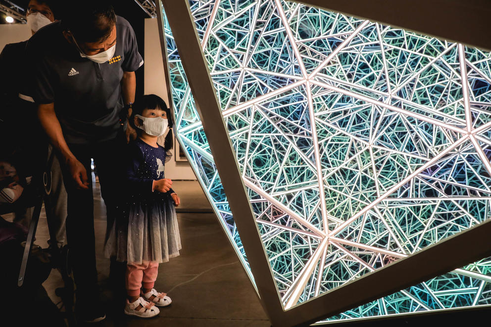 young girl with mask on stands near an artwork with neon