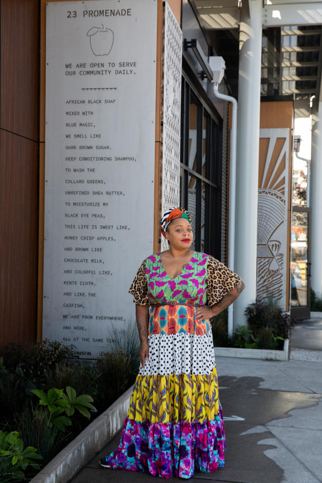 Person in headwrap and long, colorful dress in front of a stainless steel 9 foot long panel with a poem engraved in it. 