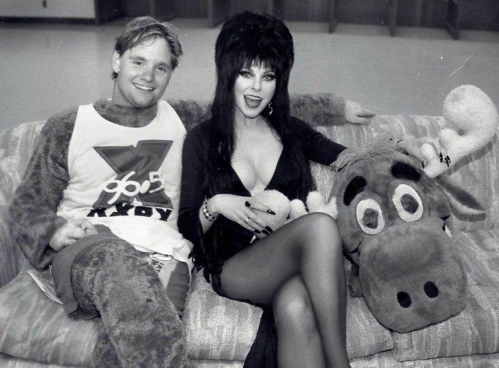 A man, in a moose suit and a woman, dresses as Elvira, sit on a couch with the stuffed moose head beside them