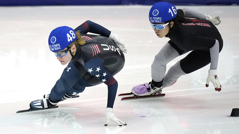 Two speedskaters in dark uniforms and blue helmets are on the ice, one following the other, both of their hands on on near the ice as they make a turn
