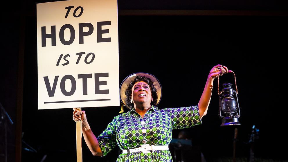 a woman in costume on stage holds a sign reading To Hope Is To Vote