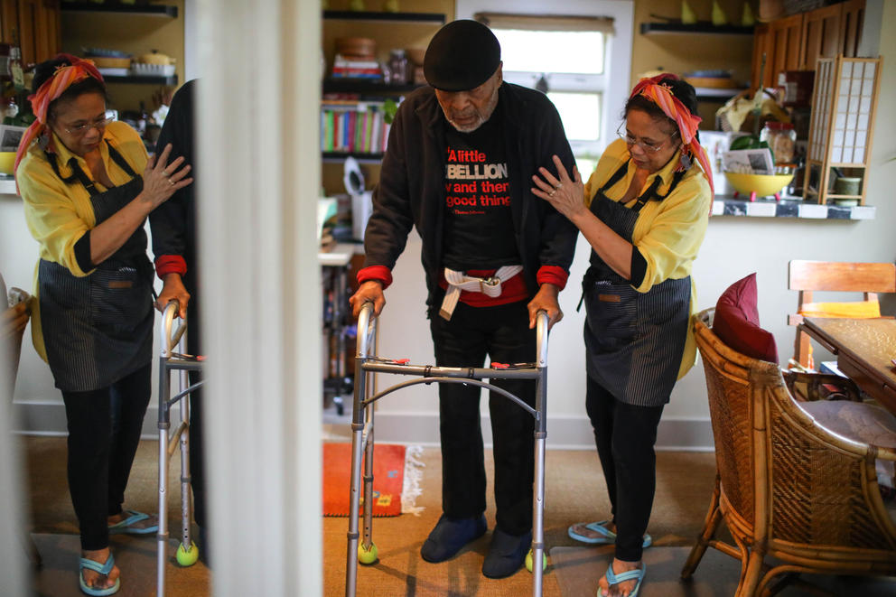A woman holds a man by the arm as he uses a walker to move around a kitchen