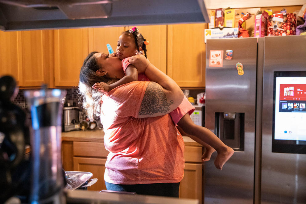 a mother holds her toddler up to give her a kiss, standing in the kitchen