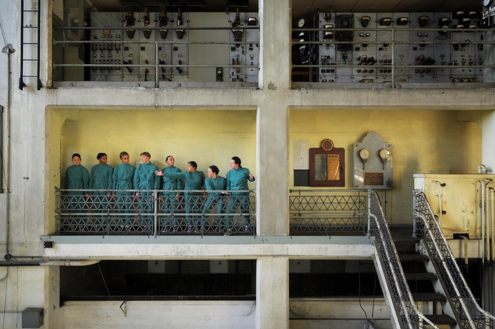 a line of dancers wearing coveralls inside a large factory