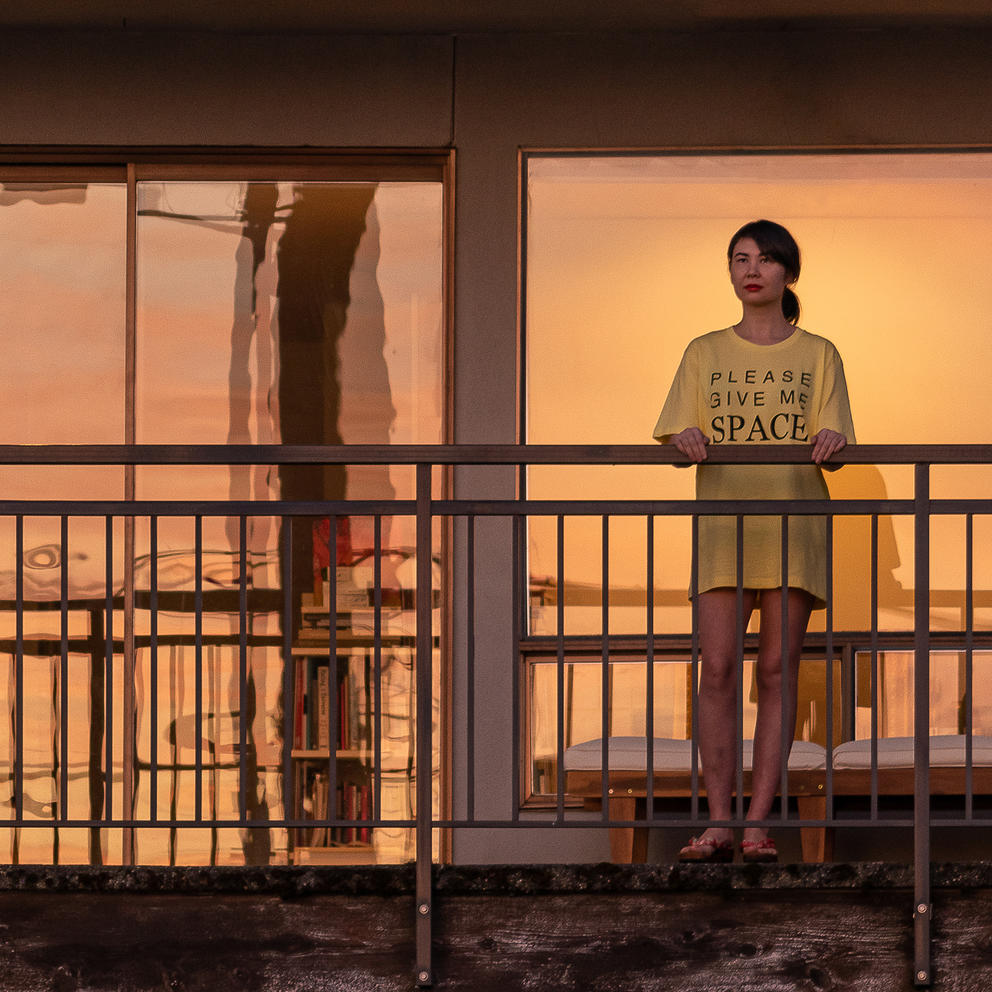 Person in yellow t-shirt saying "Please give me space" standing on a balcony, sunset reflected on the windows behind them