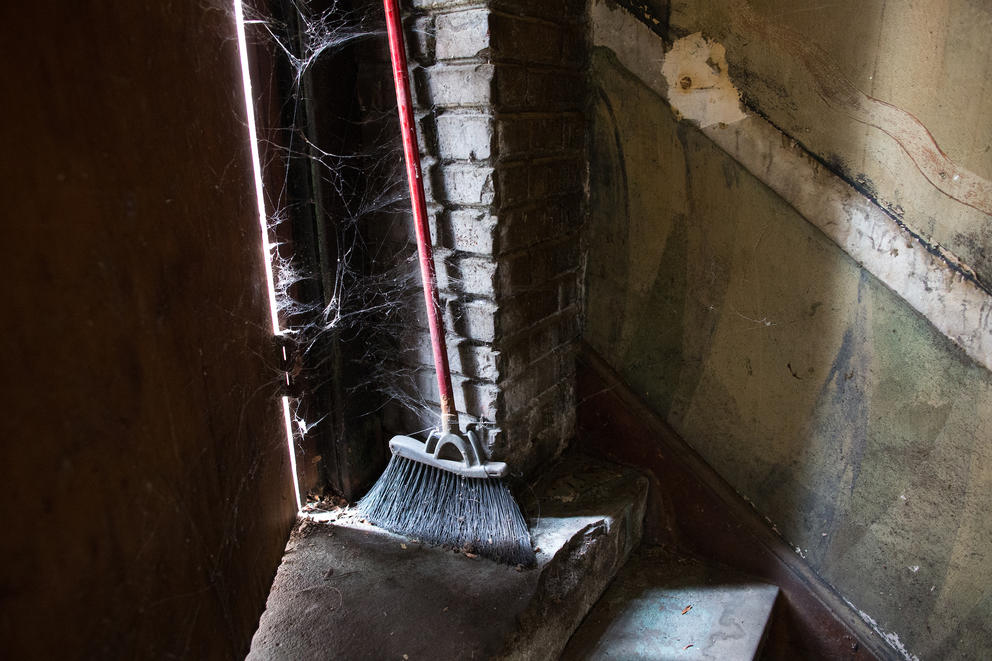 A dusty broom sits next to a doorway at the top of a staircase where old murals line the walls.