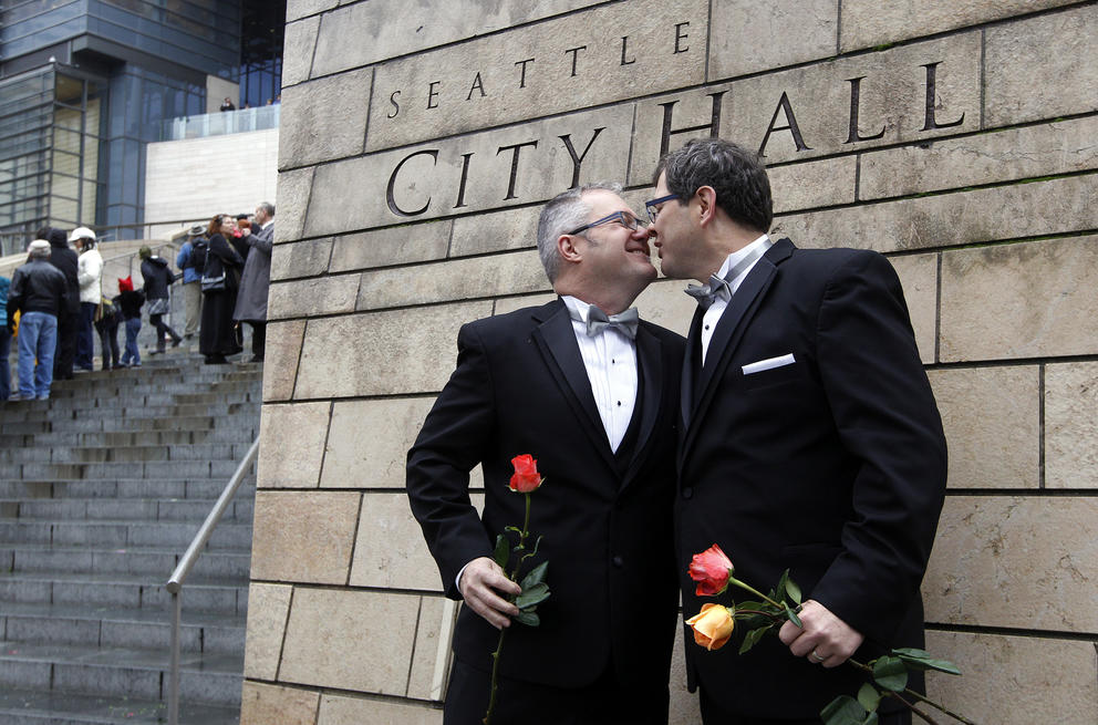 Two men in tuxedos hold flowers and kiss in front of the Seattle City Hall building
