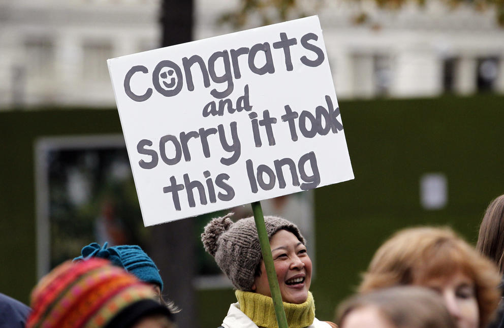 A woman in a crowd holds a sign that reads "congrats and sorry it took so long"