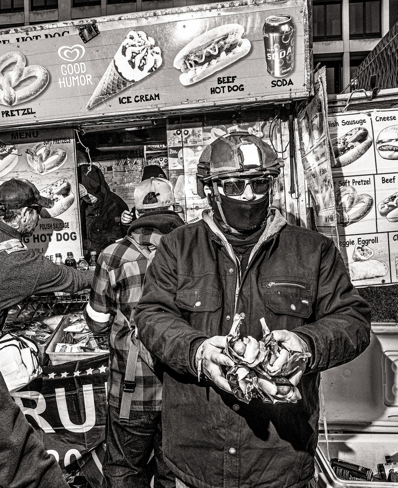Person in helmet, sunglasses and face covering carries hot dogs in his hands, in a black and white photo