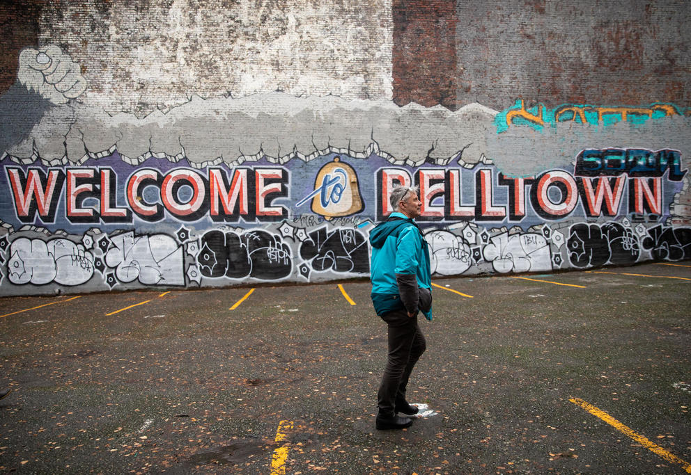 Man in blue jacket and black pants walks by mural that says "welcome to belltown." Below it are throw-ups, simple graffiti drawings