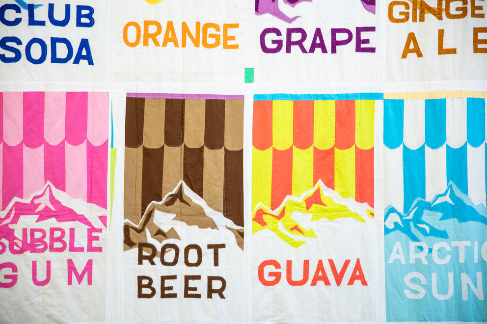 detail of a quilt featuring colorful squares with text reading: Root Beer, Guava, Grape, Orange, and more