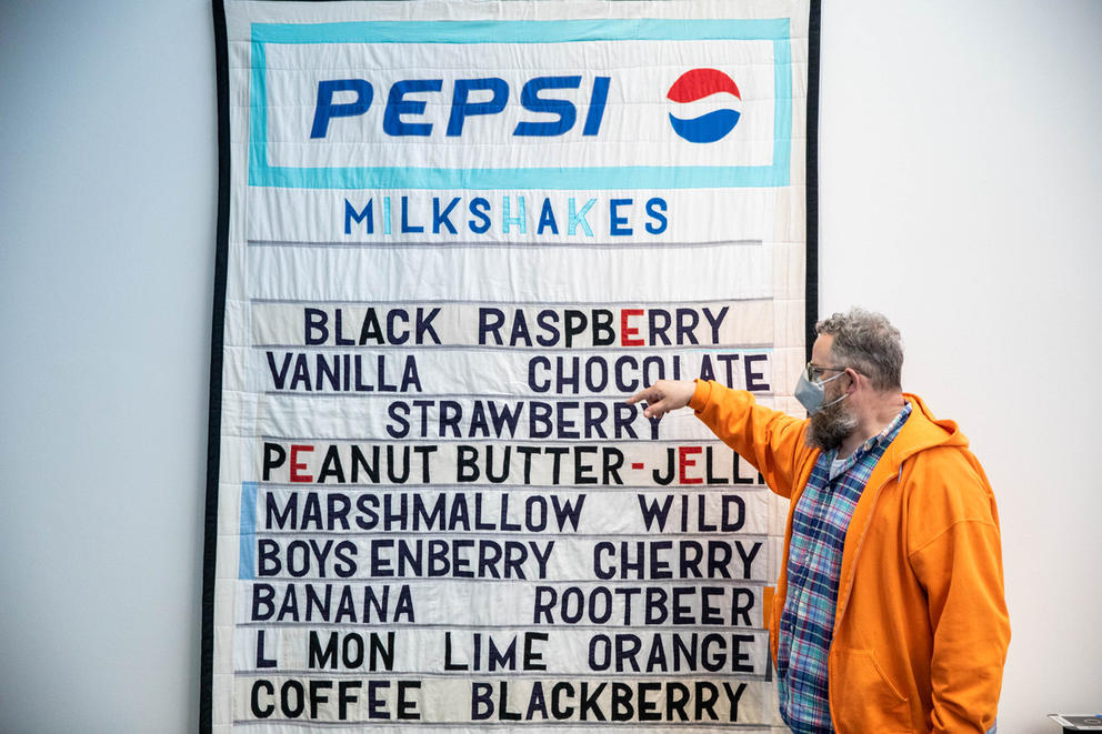 Person in plaid shirt with orange sweater pointing to a quilt hanging on the wall, featuring a sign saying PEPSI and featuring ice cream flavors