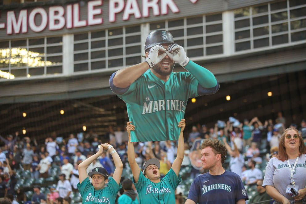 three men hold up a giant cut out of a Mariners play and cheer in a stadium