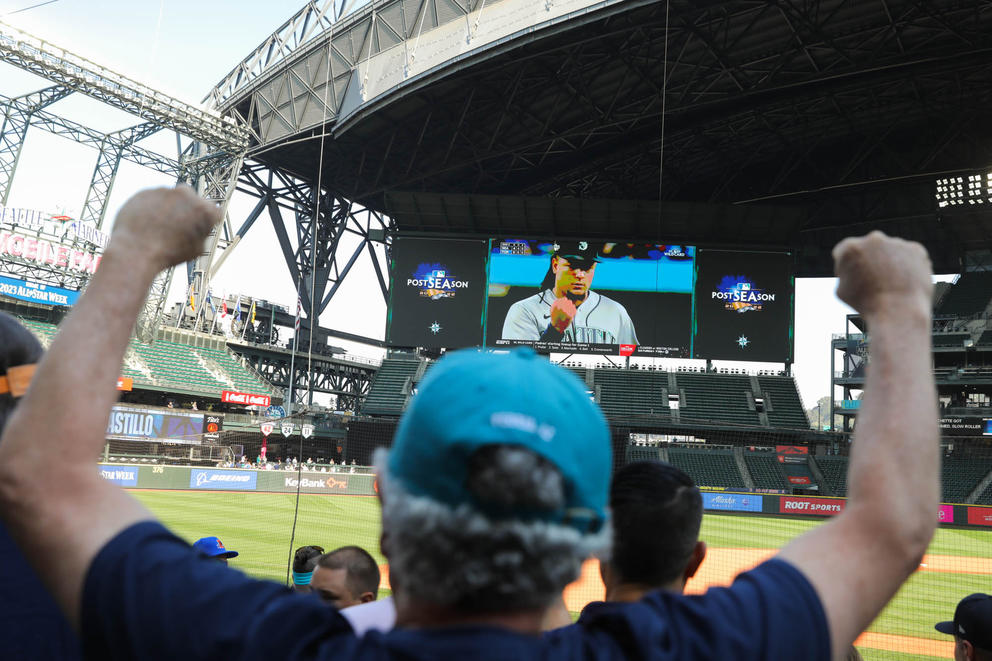 A man in the foreground holds up his fists as a baseball game plays on a giant screen in the background