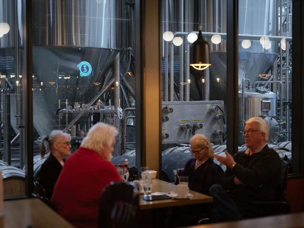 A group of people sit at a table, behind them a large window gives a view of distillery equipment