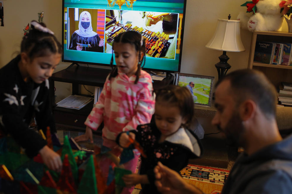 A woman in a hijab and facemask is seen on a tv behind three girls who are playing in a living room.