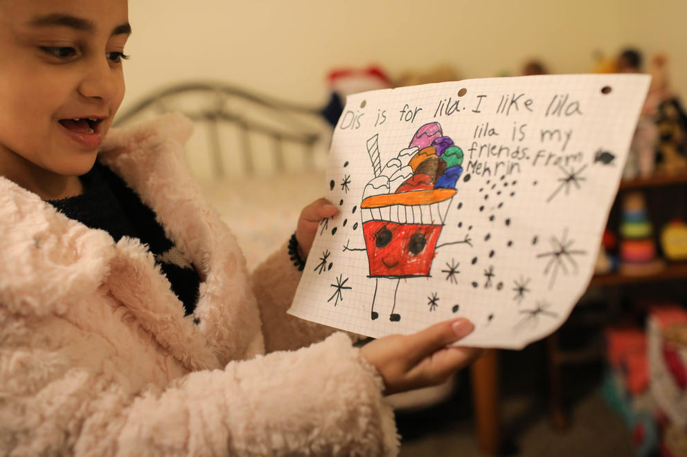 A girl holds up a drawing of multicolored ice cream. Writing reads "Dis is for lila, I like lila. lila is my friend. From Mehrin"