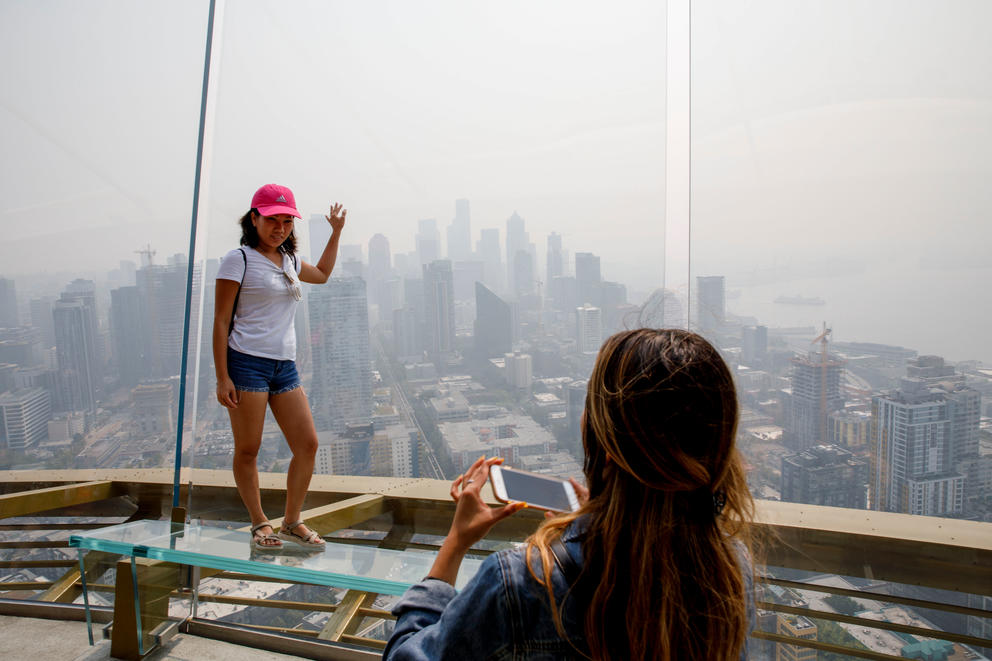 A woman poses for a photo at the top of the Space Needle. The city skyline behind her is smoky