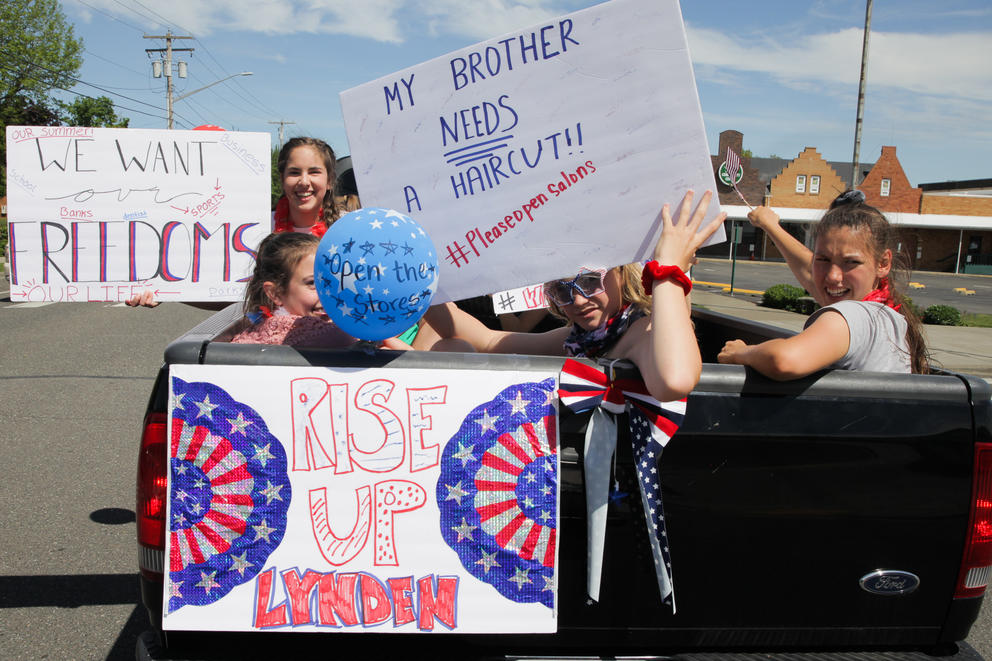 Four girls hold up protest signs ("We want our freedoms," "RISE UP," and "My brother needs a haircut!! #pleaseopensalons") while riding in a truck bed