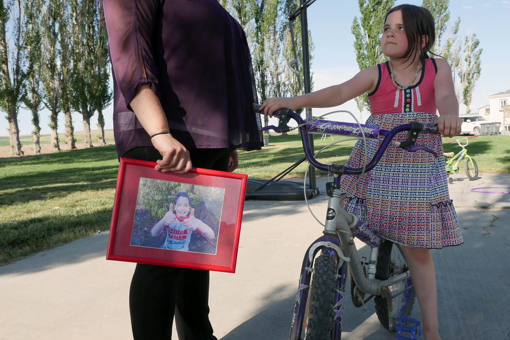 A girl on a bike looks at her mom who stands holding a framed picture of a boy