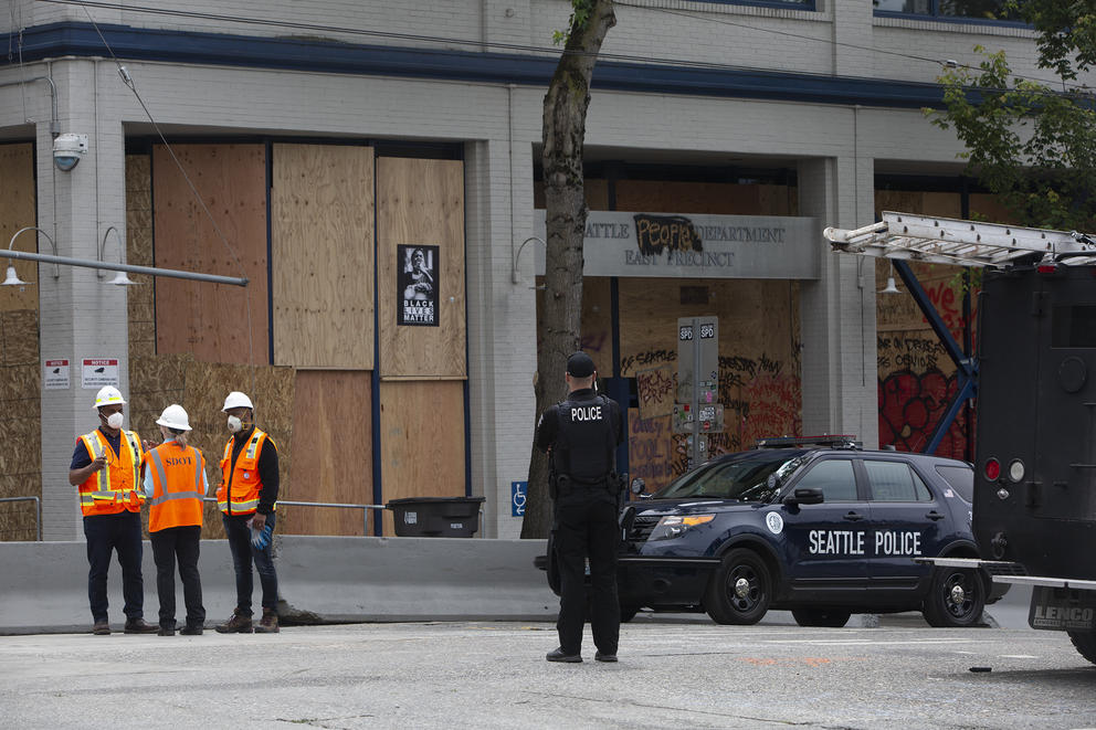 a police officer stands in middle of street, looks at a boarded up precinct with a Black Lives Matter poster on it