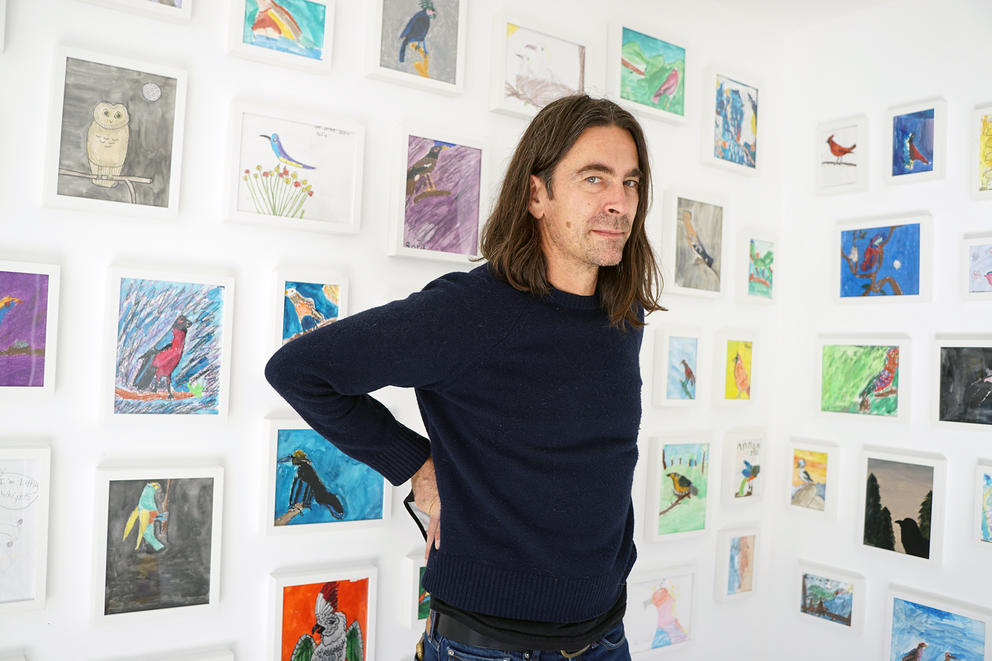 White man with long dark hair and dark t-shirt stands in a white room full of art on the walls
