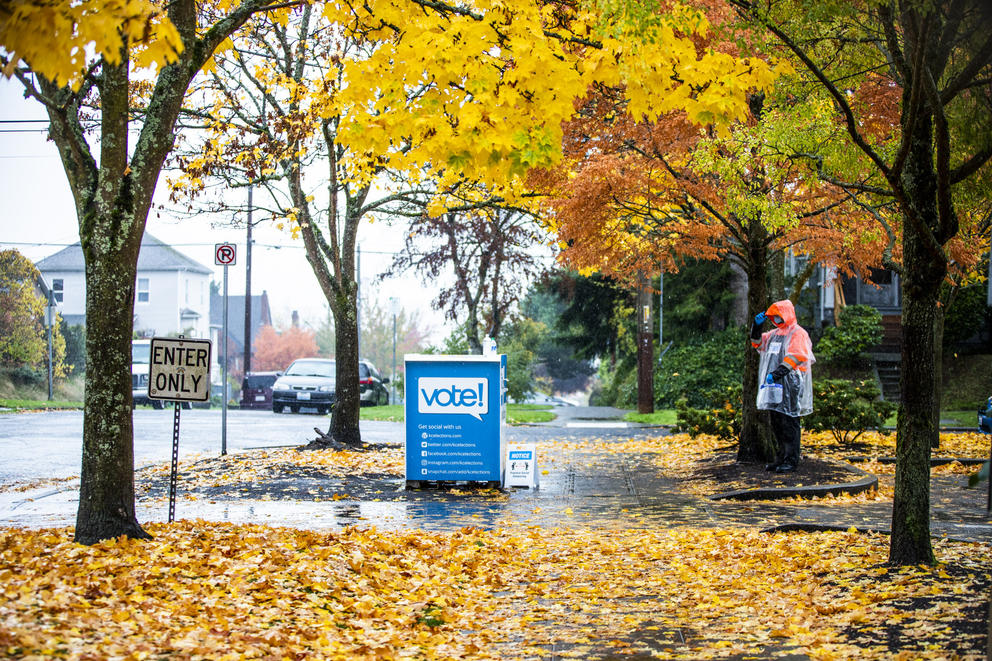 An elections worker stands near a ballot drop box among colorful fall trees