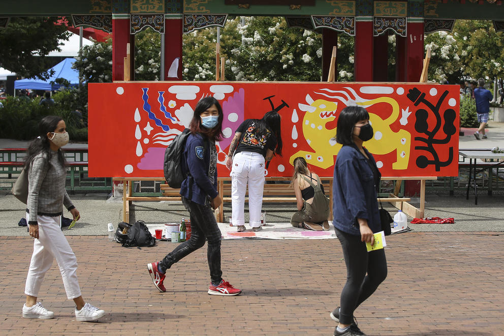 Person is painting a large wooden board in red and yellow, while people in front, in darker clothes, are walking by to the right and outside the frame