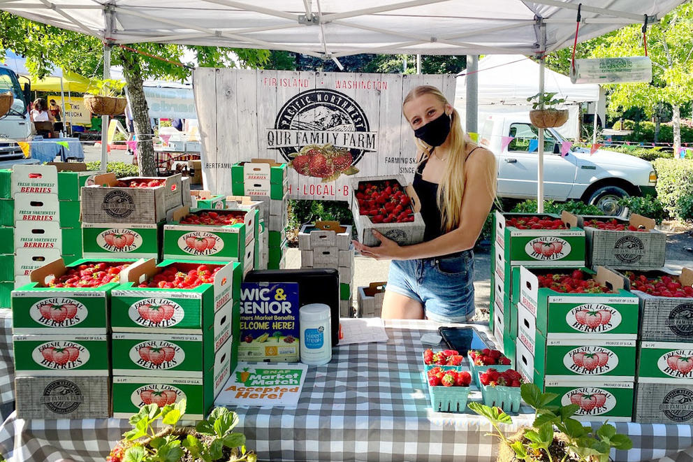 A woman holds a box of strawberries. She is surrounded by other stacked boxes of strawberries, which sit on a table with a blue checked tablecloth at a farmers market 