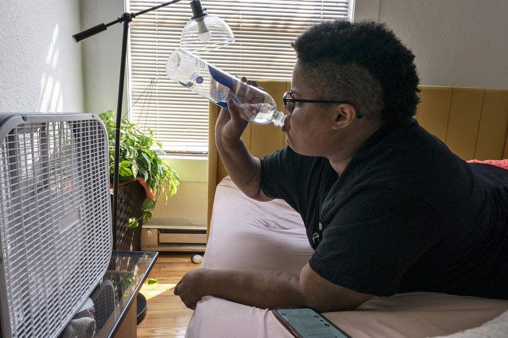 a person with short hair lies on a bed in front of a box fan drinking water out of a plastic water bottle. There is a window in the background with a shade fully drawn. 