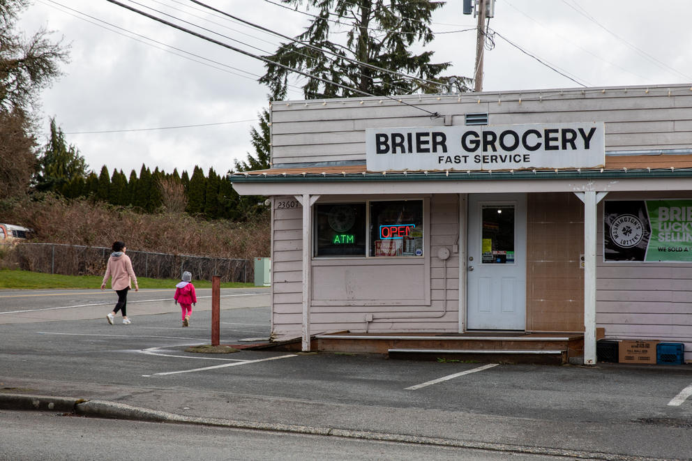 A woman in pink and a small child walk past a faded pink storefront with a sign reading "Brier Grocery, fast service."