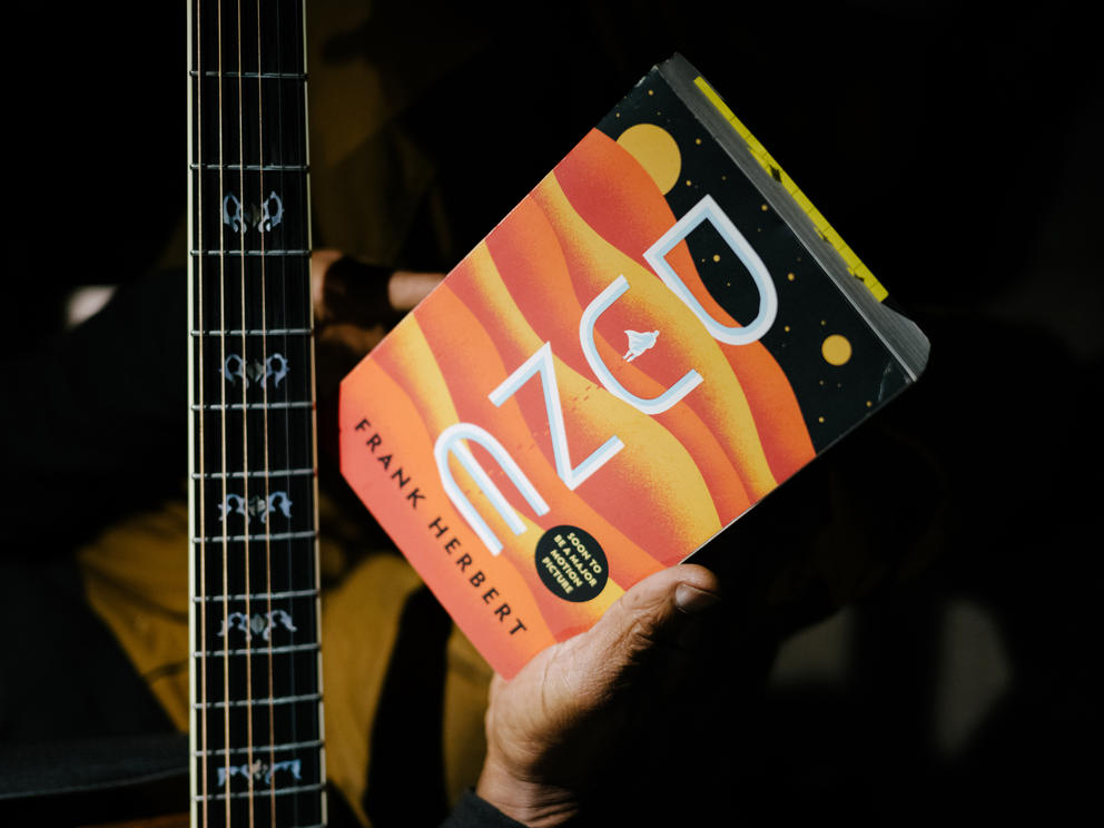 Strings of a guitar with a hand holding a book with an orange cover that states "DUNE"