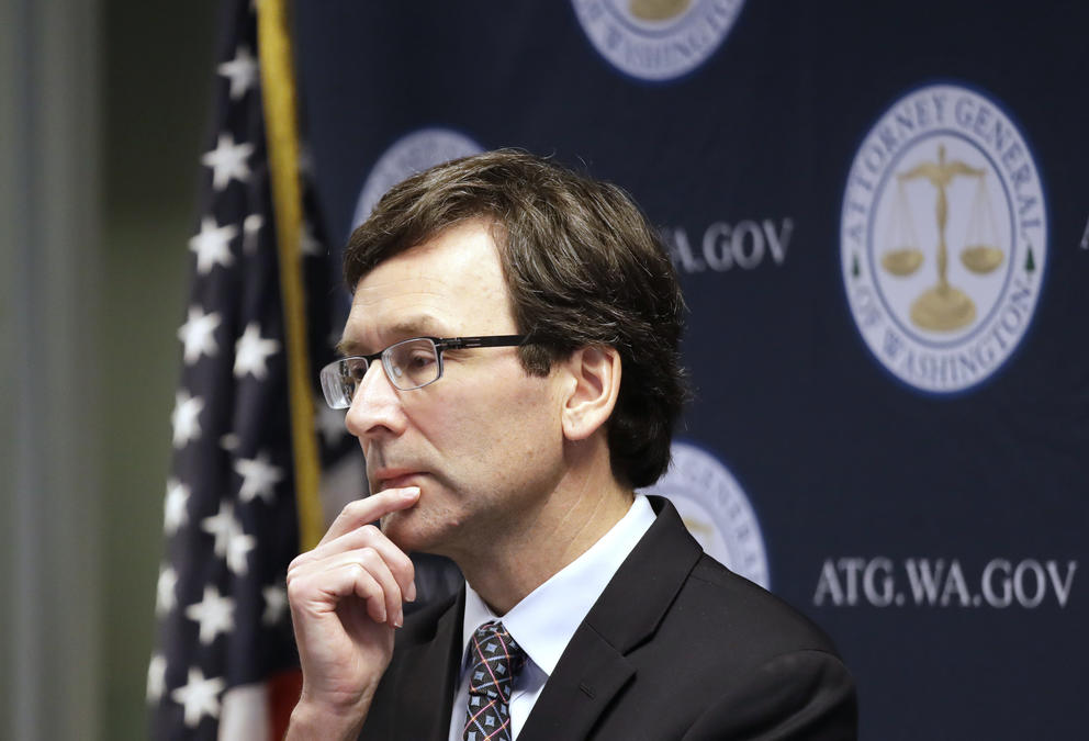A man wearing glasses and a suit faces away from the camera, holding his chin in his hand. There is a flag behind him. 
