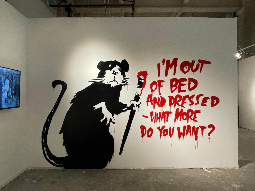 white wall featuring a large rat with a brush in its hands, with text saying: "I'm out of bed and dressed, what more do you want?"
