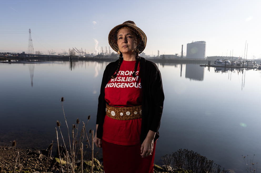 Annette Bryan stands with an industrial area's skyline behind her. Her shirt reads: "Strong, Resilient, Indigenous"