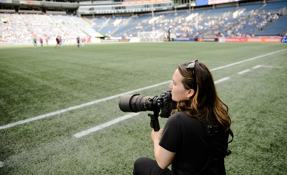 A woman holding a camera while standing on the sidelines of a soccer pitch