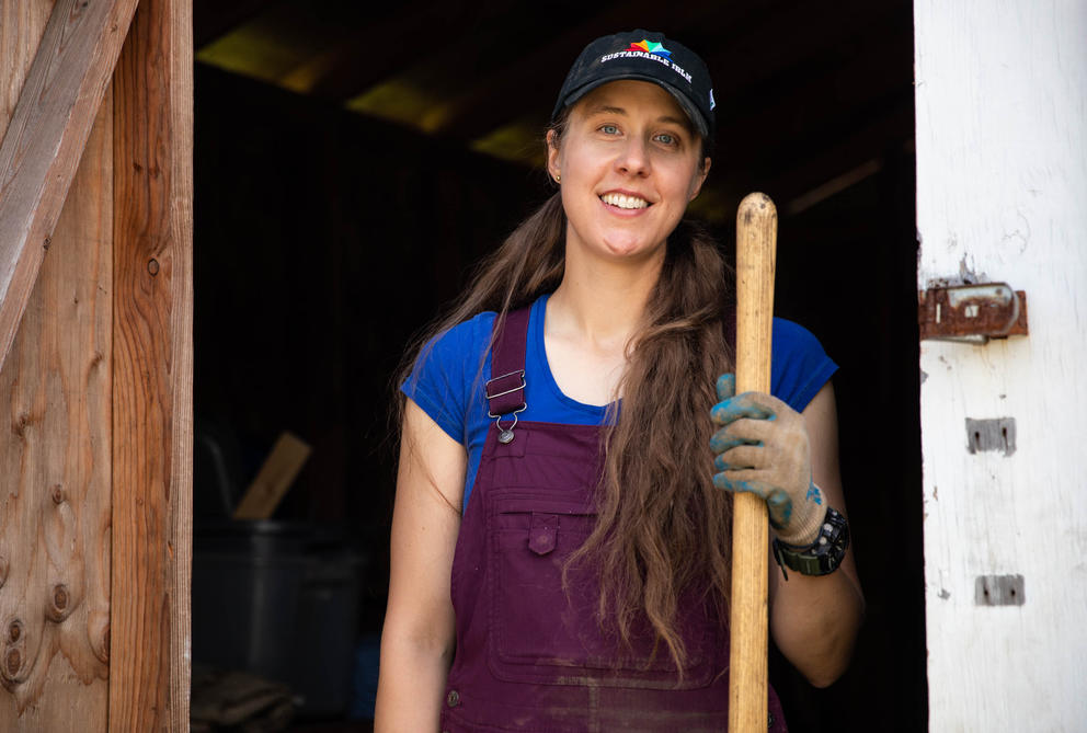 a person in purple overalls with long brown hair holds a shovel while smiling at the camera