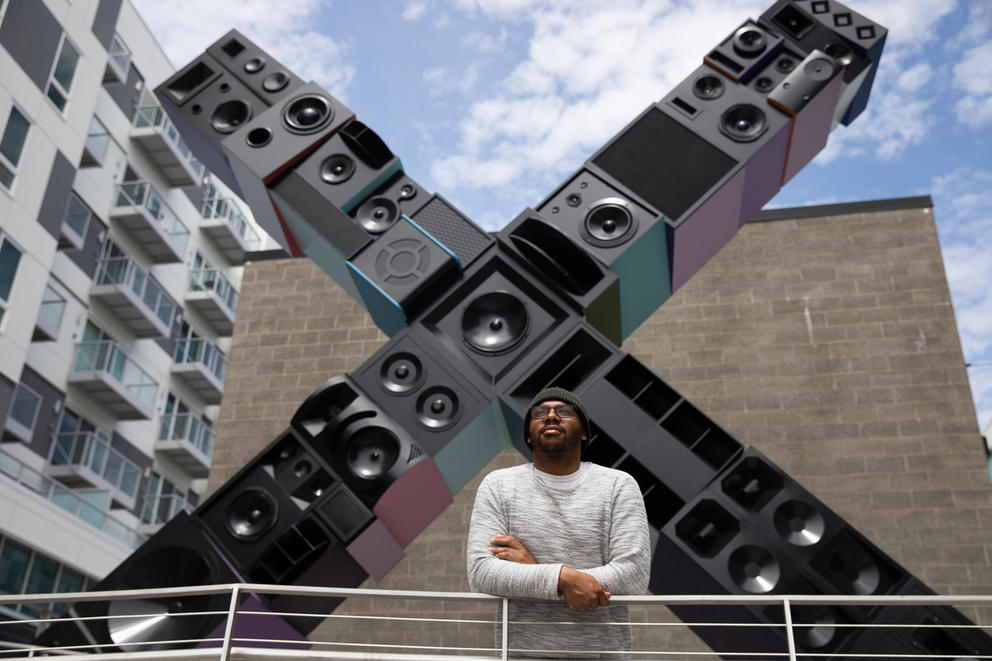 Person in hat in front of large 20 by 20 foot X sculpture made out of stacked boxes that look like speakers