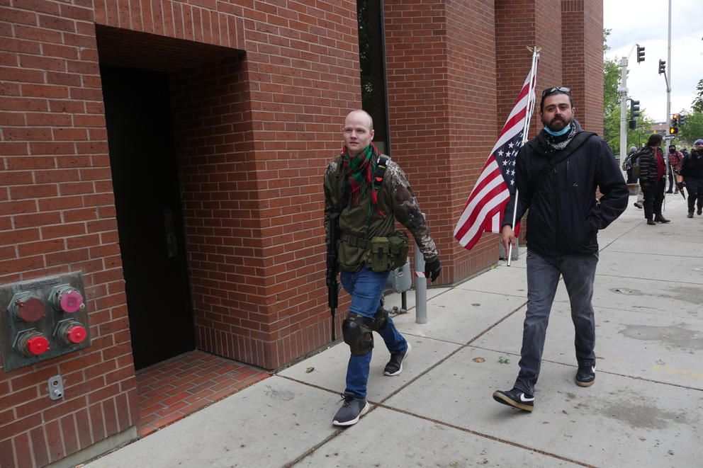A man carries an AR-15 next to another man with an upside down American flag
