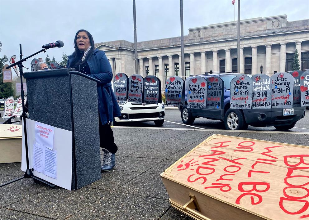 A woman stands outdoor at a podium at left, with a government building behind her. Also behind her are a string of paper signs in the shape of gravestones with names and dates on them. At right is a box shaped like a coffin that says, "Blood will be on your hands." 