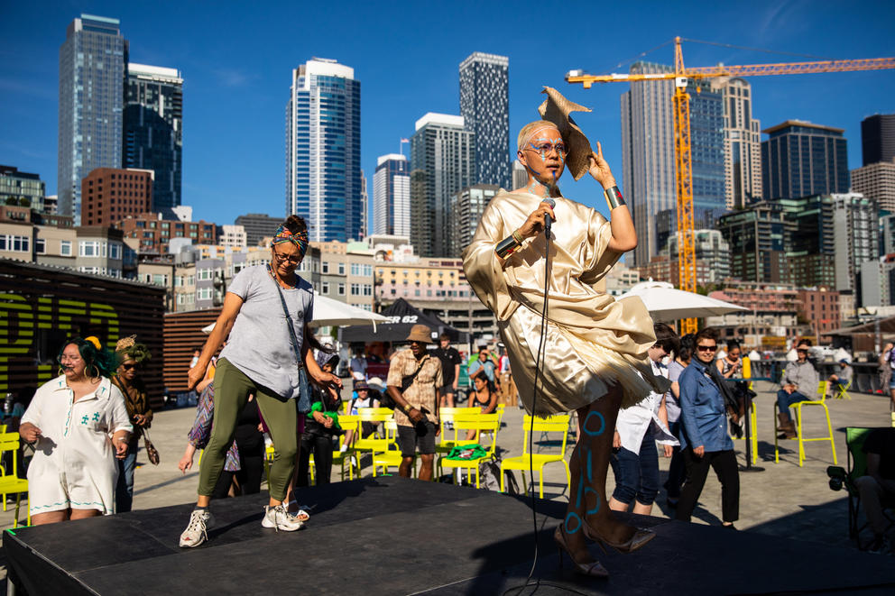 A person wearing gold holding a microphone and another person dance on a stage with the Seattle skyline behind them