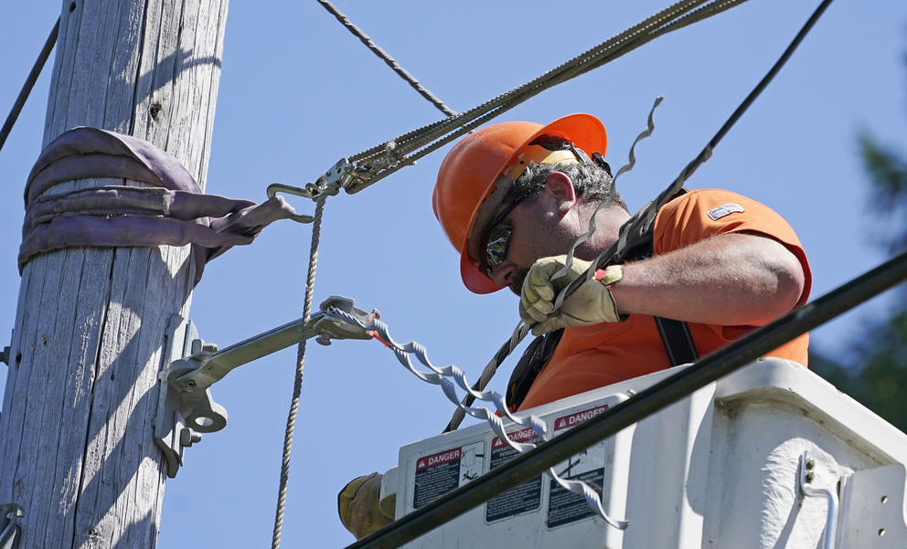 A utility worker wearing an orange shirt and helmet installs fiber cable on a utility pole while standing in a lift bucket.
